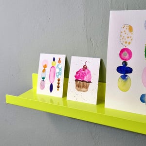 May include: Three watercolor paintings on a yellow shelf. The paintings are of a cupcake, a stack of colorful circles, and a stack of colorful ovals.
