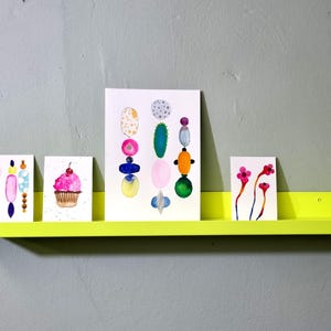 May include: Four colorful watercolor paintings on white cardstock are displayed on a bright yellow shelf. The paintings depict a cupcake, a bouquet of flowers, a geometric design, and a series of abstract shapes.