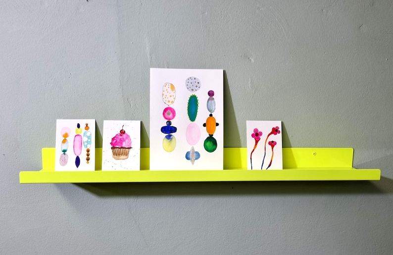May include: Four colorful watercolor paintings on white cardstock are displayed on a bright yellow shelf. The paintings depict a cupcake, a bouquet of flowers, a geometric design, and a series of abstract shapes.