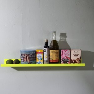 May include: A yellow shelf with a variety of items on it, including two avocados, a can of cocoa powder, a can of salt, a bottle of oil, a bottle of vinegar, and a box of vanilla extract. The shelf is mounted on a white wall.