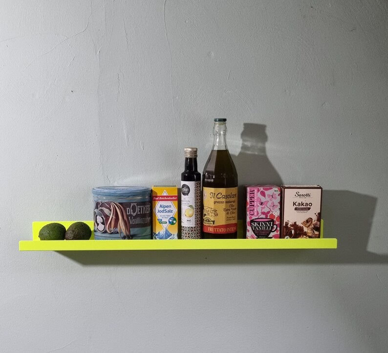May include: A yellow shelf with a variety of items on it, including two avocados, a can of cocoa powder, a can of salt, a bottle of oil, a bottle of vinegar, and a box of vanilla extract. The shelf is mounted on a white wall.