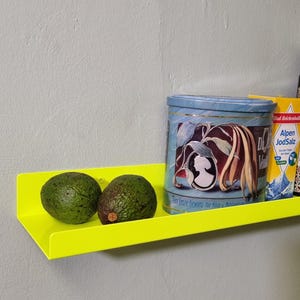 May include: A bright yellow metal shelf with two green avocados on top. The shelf is mounted on a white wall. A blue and white tin can with the text "Das Beste Gew&uuml;rz f&uuml;r Milch & Mehlspeisen" is on the shelf. A blue and white box with the text "Echt Reichenhaller Alpen JodSalz" is on the shelf.