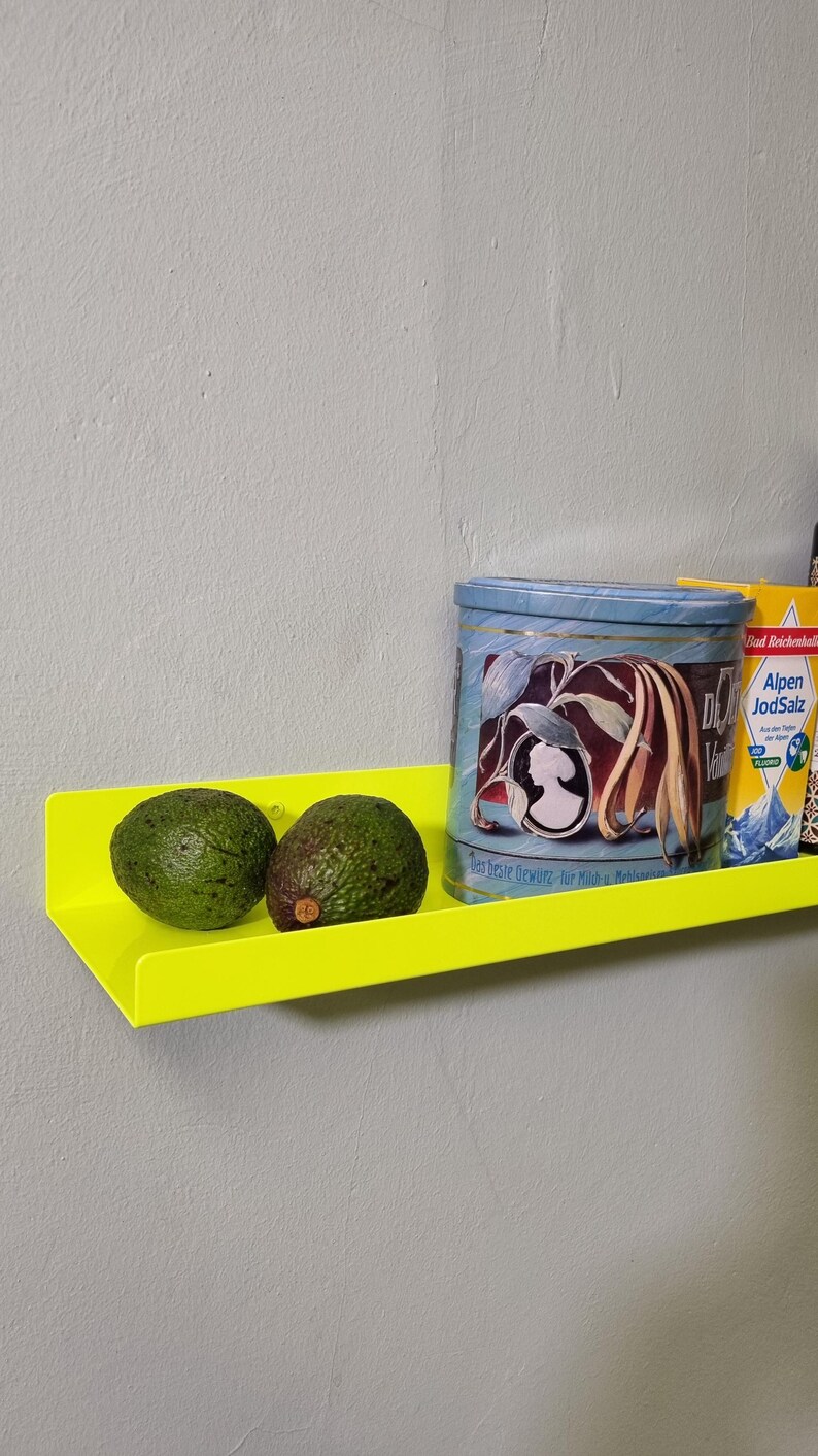 May include: A bright yellow metal shelf with two green avocados on top. The shelf is mounted on a white wall. A blue and white tin can with the text "Das Beste Gew&uuml;rz f&uuml;r Milch & Mehlspeisen" is on the shelf. A blue and white box with the text "Echt Reichenhaller Alpen JodSalz" is on the shelf.