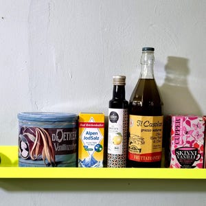 May include: A yellow shelf with a variety of items on it, including a can of vanilla extract, a box of Alpen JodSalz, a bottle of olive oil labeled "Il Casolare", a box of tea labeled "Skinni Vanilli", a box of cocoa labeled "Sanetti Kakao", and two avocados.