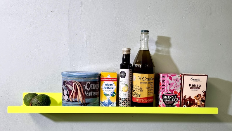 May include: A yellow shelf with a variety of items on it, including a can of vanilla extract, a box of Alpen JodSalz, a bottle of olive oil labeled "Il Casolare", a box of tea labeled "Skinni Vanilli", a box of cocoa labeled "Sanetti Kakao", and two avocados.