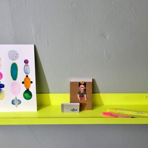 May include: A bright yellow metal shelf with rounded edges, holding a print of colorful abstract shapes, a small notebook with a cartoon image, a highlighter, and a pen.