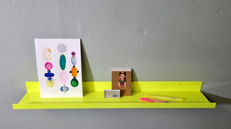 May include: A bright yellow metal shelf with rounded edges, holding a print of colorful abstract shapes, a small notebook with a cartoon image, a highlighter, and a pen.