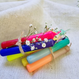 Könnte beinhalten: Ein Bündel bunter Geburtstagskerzen, die mit Schnur zusammengebunden und mit kleinen weißen Blüten verziert sind. Die Kerzen sind in den Farben Rot, Blau, Gelb, Grün, Lila und Orange.