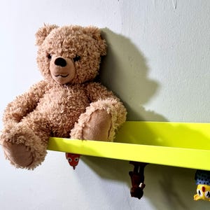 May include: A brown teddy bear sits on a bright yellow shelf with three colorful toy figures hanging from the bottom.
