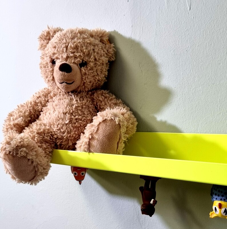 May include: A brown teddy bear sits on a bright yellow shelf with three colorful toy figures hanging from the bottom.