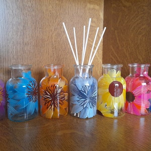May include: Seven clear glass vases, each hand-painted with a different colourful floral design. One vase contains white diffuser sticks. The flowers are in shades of pink, blue, orange, yellow, and red.
