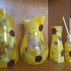 May include: Four glass vases with hand-painted sunflowers in shades of yellow and brown. The vases are arranged in a row on a wooden shelf.
