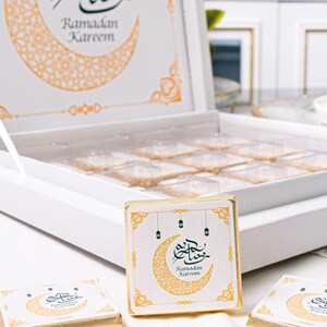 Op de afbeelding: Een witte doos met een doorzichtig deksel, met individueel verpakte traktaties. De doos is versierd met een maan en de tekst "Ramadan Kareem". Drie verpakte traktaties liggen ervoor, elk met hetzelfde ontwerp.