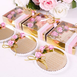 Peut inclure: Quatre petites bo&icirc;tes cadeaux dor&eacute;es avec un ruban rose et blanc et un dessus transparent. Chaque bo&icirc;te contient un petit bouquet de fleurs roses. Trois plateaux ronds, blancs et d&eacute;coratifs avec une surface miroir dor&eacute;e et des fleurs roses. Les plateaux ont du texte arabe sur la surface du miroir.