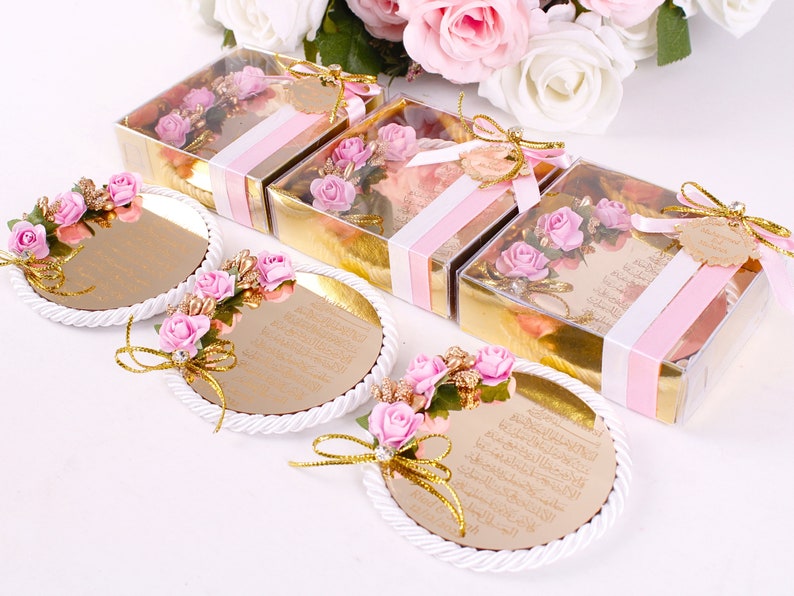 Peut inclure: Quatre petites bo&icirc;tes cadeaux dor&eacute;es avec un ruban rose et blanc et un dessus transparent. Chaque bo&icirc;te contient un petit bouquet de fleurs roses. Trois plateaux ronds, blancs et d&eacute;coratifs avec une surface miroir dor&eacute;e et des fleurs roses. Les plateaux ont du texte arabe sur la surface du miroir.
