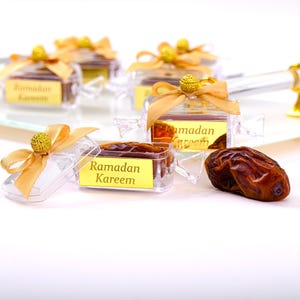 Puede incluir: Cajas de plástico transparente con cinta dorada y el texto "Ramadan Kareem" llenas de dátiles. Las cajas están sobre una superficie blanca.