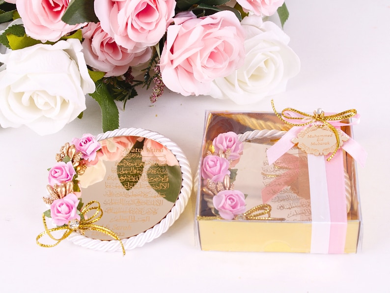 Peut inclure: Une bo&icirc;te ronde blanche avec un design de corde dor&eacute;e et un motif floral rose et or. La bo&icirc;te contient un miroir rond avec une inscription dor&eacute;e en arabe. La bo&icirc;te est attach&eacute;e avec un ruban rose et une &eacute;tiquette dor&eacute;e qui dit "Muhammad Eid Mubarak".