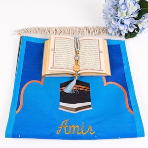 May include: A blue prayer rug with a Kaaba design and the name "Amir" embroidered in gold. An open Quran and a beaded prayer accessory rest on the rug. A bouquet of blue flowers is in the background.
