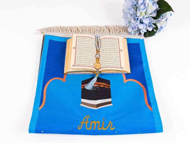 May include: A blue prayer rug with a Kaaba design and the name "Amir" embroidered in gold. An open Quran and a beaded prayer accessory rest on the rug. A bouquet of blue flowers is in the background.