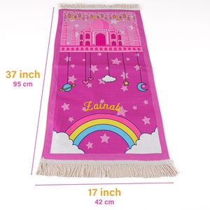 Puede incluir: Alfombra de oraci&oacute;n rosa con un arco&iacute;ris, estrellas y el nombre "Lainab" impreso en ella. La alfombra mide 95 cm por 42 cm.
