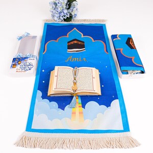 May include: A blue prayer rug with a Mecca design, a Quran, and a gift box. The rug features a cloud and rainbow design with the name "Amir". The gift box has "Eid Mubarak" text.