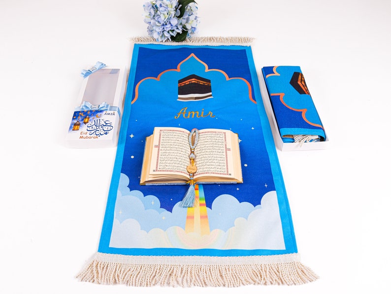 May include: A blue prayer rug with a Mecca design, a Quran, and a gift box. The rug features a cloud and rainbow design with the name "Amir". The gift box has "Eid Mubarak" text.