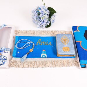 May include: A blue prayer mat, a Quran, and a prayer bead set are displayed. The mat and Quran are personalized with the name "Amir". The set includes a gift box with "Eid Mubarak" text and a blue tassel.