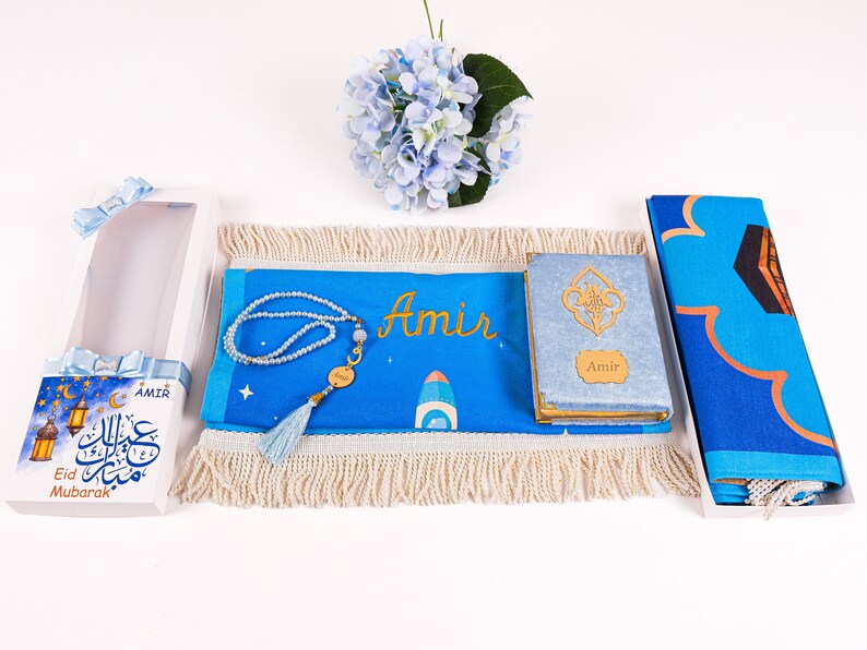 May include: A blue prayer mat, a Quran, and a prayer bead set are displayed. The mat and Quran are personalized with the name "Amir". The set includes a gift box with "Eid Mubarak" text and a blue tassel.