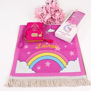 Puede incluir: Alfombra de oraci&oacute;n rosa con un dise&ntilde;o de arco&iacute;ris y estrellas. La alfombra tiene un borde de flecos blancos y el nombre "Zainab" est&aacute; bordado en ella. Se incluye un libro de oraci&oacute;n de terciopelo rosa con un borla a juego y un juego de cuentas de oraci&oacute;n.