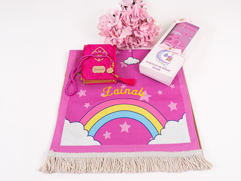 Puede incluir: Alfombra de oraci&oacute;n rosa con un dise&ntilde;o de arco&iacute;ris y estrellas. La alfombra tiene un borde de flecos blancos y el nombre "Zainab" est&aacute; bordado en ella. Se incluye un libro de oraci&oacute;n de terciopelo rosa con un borla a juego y un juego de cuentas de oraci&oacute;n.