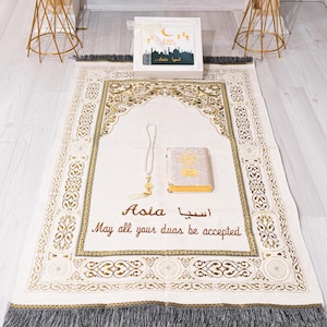 Puede incluir: Una alfombra de oración crema con un borde dorado decorativo y flecos grises. La alfombra tiene la palabra "Asia" en inglés y árabe. Un pequeño libro, un collar y una caja de regalo están sobre la alfombra. El texto "May all your duas be accepted" también está presente.