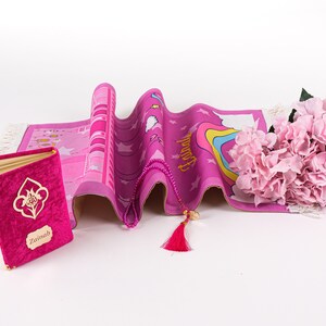 Puede incluir: Alfombra de oraci&oacute;n rosa con un arco&iacute;ris, estrellas y la palabra "Hamd" impresa en ella. Tambi&eacute;n se incluye un libro de oraci&oacute;n rosa con el nombre "Zainab" y un collar de oraci&oacute;n de cuentas rosas.