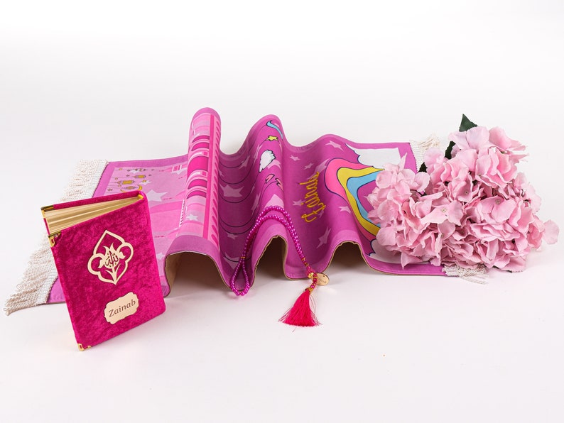 Puede incluir: Alfombra de oraci&oacute;n rosa con un arco&iacute;ris, estrellas y la palabra "Hamd" impresa en ella. Tambi&eacute;n se incluye un libro de oraci&oacute;n rosa con el nombre "Zainab" y un collar de oraci&oacute;n de cuentas rosas.