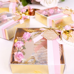 Peut inclure: Une petite bo&icirc;te cadeau dor&eacute;e avec un couvercle transparent. La bo&icirc;te contient un ornement rond en miroir avec des fleurs roses et des accents dor&eacute;s. L'ornement est orn&eacute; d'un ruban rose et d'une &eacute;tiquette qui dit "Mohammed Eid Mubarak".