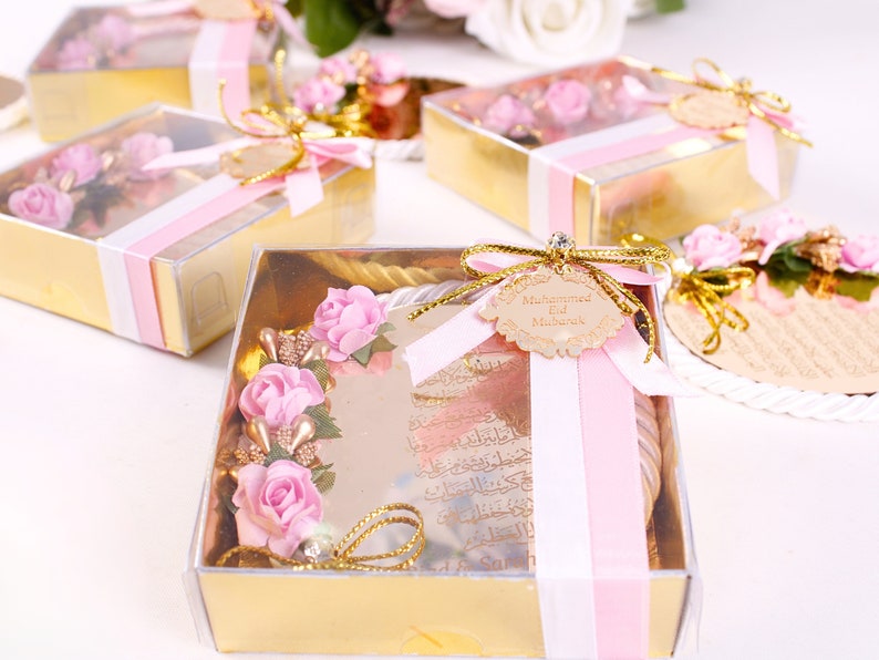 Peut inclure: Une petite bo&icirc;te cadeau dor&eacute;e avec un couvercle transparent. La bo&icirc;te contient un ornement rond en miroir avec des fleurs roses et des accents dor&eacute;s. L'ornement est orn&eacute; d'un ruban rose et d'une &eacute;tiquette qui dit "Mohammed Eid Mubarak".