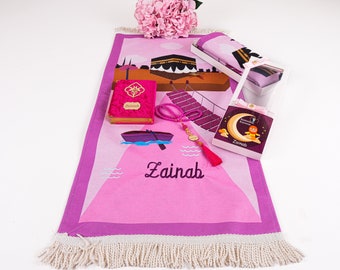 May include: Pink prayer rug with a white fringe, a pink prayer book with the name "Zainab" on the cover, a pink prayer bead necklace, a small pink boat, and a pink box with a crescent moon and the name "Zainab" on it.