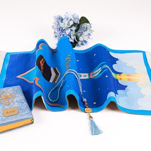 May include: A blue prayer rug with a colorful design, a blue beaded rosary, and a blue book with gold accents. The rug features a unique pattern with a star and cloud motif. A bouquet of blue flowers is in the background.