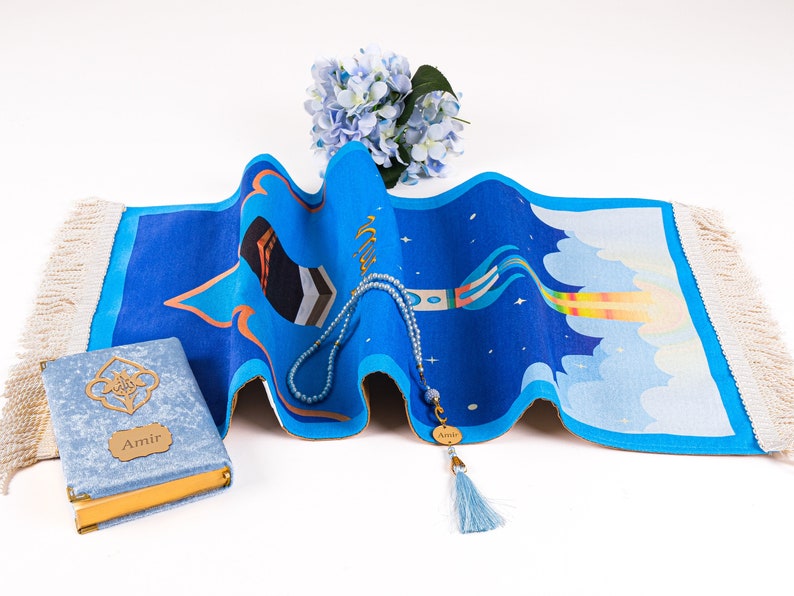 May include: A blue prayer rug with a colorful design, a blue beaded rosary, and a blue book with gold accents. The rug features a unique pattern with a star and cloud motif. A bouquet of blue flowers is in the background.
