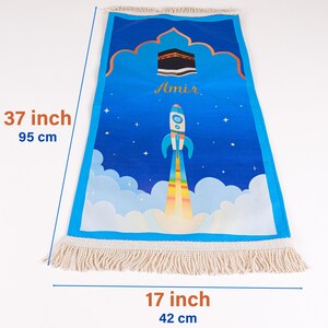 May include: Blue prayer rug, 37 inches long and 17 inches wide, with a rocket ship design. The rug features a Kaaba illustration and the name "Amir". It has a fringe border and a blue sky background.