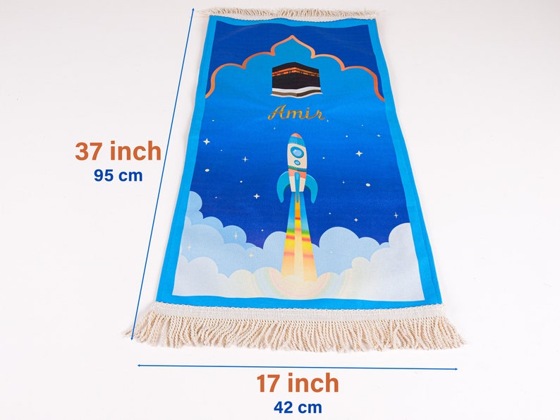 May include: Blue prayer rug, 37 inches long and 17 inches wide, with a rocket ship design. The rug features a Kaaba illustration and the name "Amir". It has a fringe border and a blue sky background.