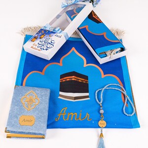 May include: A blue prayer rug with a gold arched design and a depiction of the Kaaba. The set includes a matching prayer cloth, a prayer bead necklace, and a small book. The text "Eid Mubarak" is on a gift box.