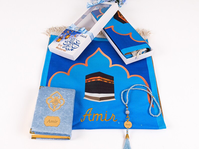May include: A blue prayer rug with a gold arched design and a depiction of the Kaaba. The set includes a matching prayer cloth, a prayer bead necklace, and a small book. The text "Eid Mubarak" is on a gift box.