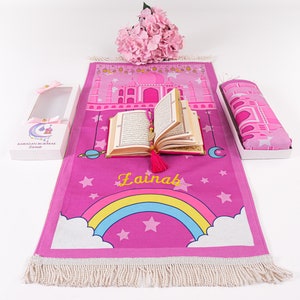 Puede incluir: Alfombra de oraci&oacute;n rosa con un arco&iacute;ris, estrellas y el nombre "Lainab" impreso en ella. La alfombra est&aacute; enrollada y viene con una alfombra de oraci&oacute;n a juego y un peque&ntilde;o libro.