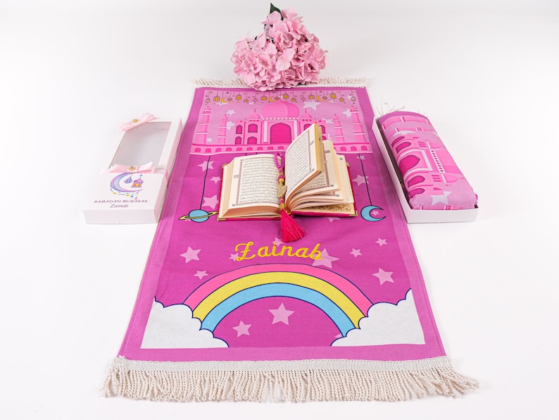 Puede incluir: Alfombra de oraci&oacute;n rosa con un arco&iacute;ris, estrellas y el nombre "Lainab" impreso en ella. La alfombra est&aacute; enrollada y viene con una alfombra de oraci&oacute;n a juego y un peque&ntilde;o libro.