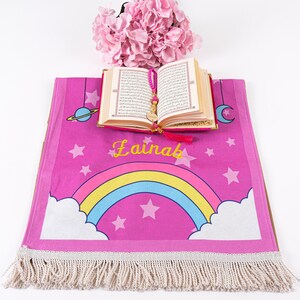 Puede incluir: Una alfombra de oraci&oacute;n rosa con un arco&iacute;ris, estrellas y el nombre "Lainab" bordado. Un peque&ntilde;o libro abierto con una borla rosa est&aacute; encima de la alfombra.