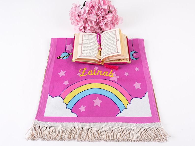 Puede incluir: Una alfombra de oraci&oacute;n rosa con un arco&iacute;ris, estrellas y el nombre "Lainab" bordado. Un peque&ntilde;o libro abierto con una borla rosa est&aacute; encima de la alfombra.
