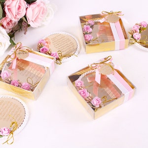 Peut inclure: Quatre petites bo&icirc;tes cadeaux avec des couvercles transparents et des finitions dor&eacute;es. Chaque bo&icirc;te contient un miroir rond, blanc et d&eacute;coratif avec des fleurs roses et des accents dor&eacute;s. Les miroirs portent du texte arabe. Les bo&icirc;tes sont attach&eacute;es avec un ruban rose et blanc.