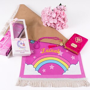Puede incluir: Una alfombra de oraci&oacute;n rosa con un dise&ntilde;o de arco&iacute;ris y estrellas. La alfombra tiene flecos blancos y el nombre "Zainab" est&aacute; escrito en ella. El conjunto tambi&eacute;n incluye una alfombra de oraci&oacute;n rosa, un libro de oraci&oacute;n de terciopelo rosa con el nombre "Zainab" en &eacute;l, un juego de cuentas de oraci&oacute;n rosa y una caja de regalo blanca que contiene una alfombra de oraci&oacute;n rosa.