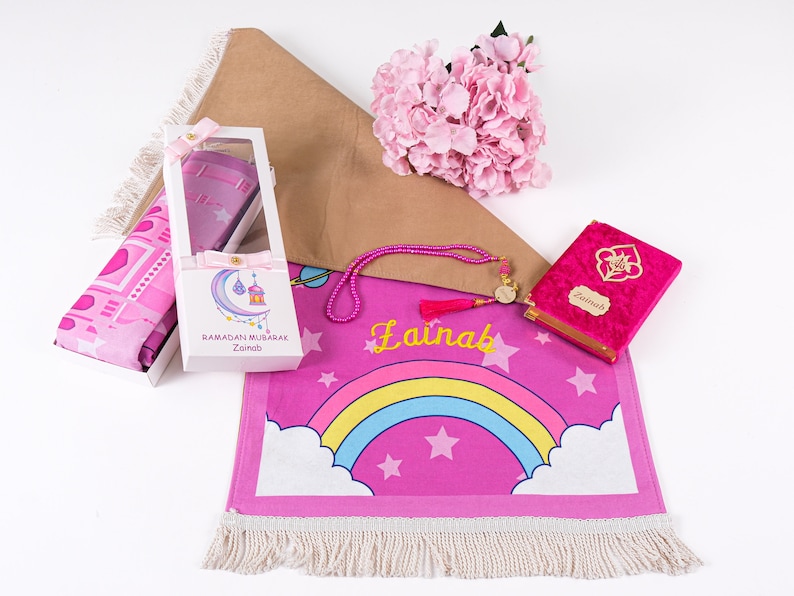 Puede incluir: Una alfombra de oraci&oacute;n rosa con un dise&ntilde;o de arco&iacute;ris y estrellas. La alfombra tiene flecos blancos y el nombre "Zainab" est&aacute; escrito en ella. El conjunto tambi&eacute;n incluye una alfombra de oraci&oacute;n rosa, un libro de oraci&oacute;n de terciopelo rosa con el nombre "Zainab" en &eacute;l, un juego de cuentas de oraci&oacute;n rosa y una caja de regalo blanca que contiene una alfombra de oraci&oacute;n rosa.