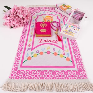 May include: Pink prayer rug with a white border and a floral design. The rug has the name "Lainah" embroidered on it. The rug is accompanied by a pink prayer book, a set of prayer beads, and a small white box with a pink ribbon.